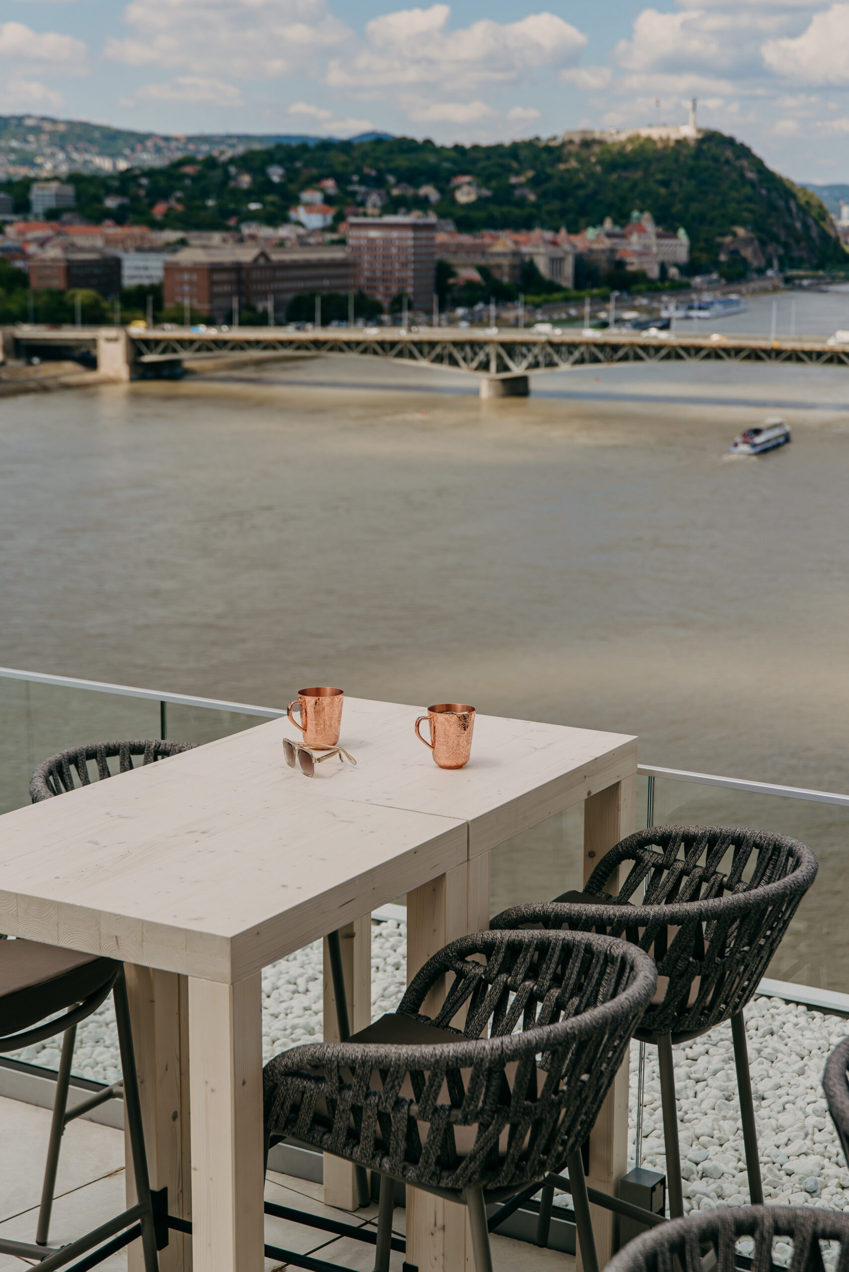 asztal réz bögrékkel és napszemüveggel, háttérben a Duna és a budapesti hidak látványa a Selva Skybar teraszáról